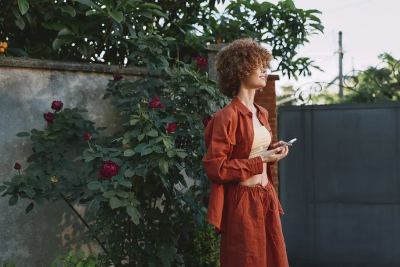 A woman with curly hair in a rust jumpsuit stands by a rose-covered wall, looking to the side with a phone in hand, capturing a casual, reflective moment