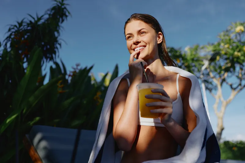 Smiling woman in swimwear wrapped in a towel holding a glass of orange juice outdoors. Sunny day with blue sky and tropical plants in the background. Concept of lifestyle and relaxation.