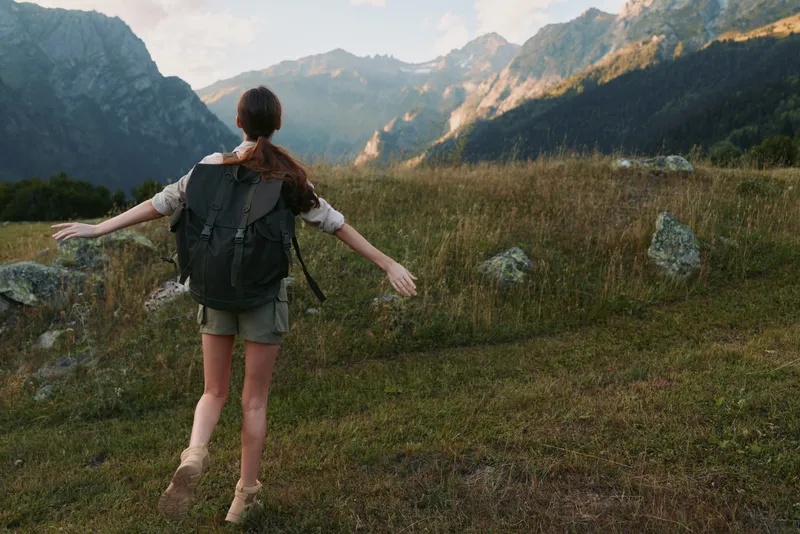 Young woman with a backpack strolls through a mountain meadow with arms outstretched, feeling carefree and free as distant peaks rise under a clear sky.