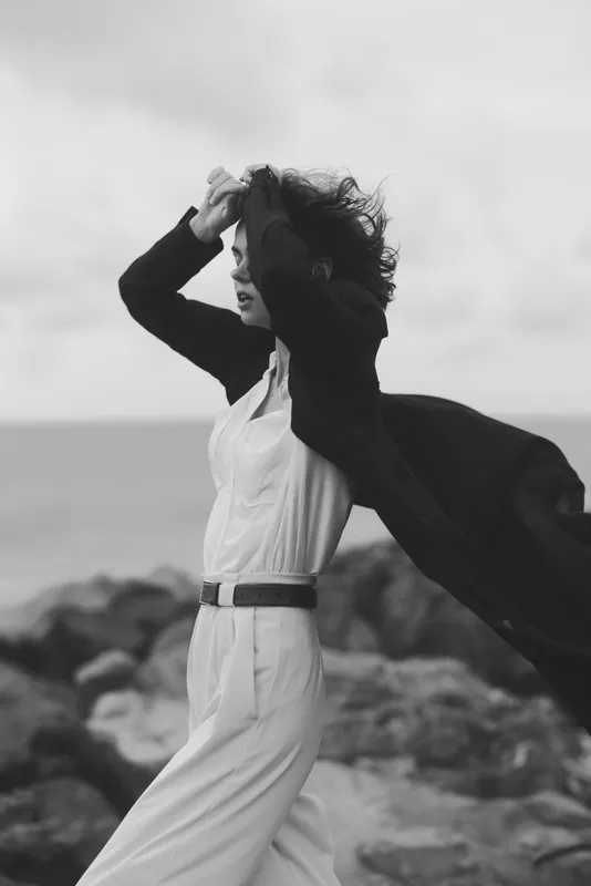 Dynamic black and white scene of a woman in a flowing white dress and coat, wind sweeping her hair by the sea, capturing movement and fashion.