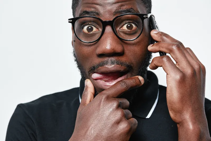 African man with dark skin and glasses talking on phone with surprised expression, wearing black shirt on white background. Closeup, communication and emotion concept.