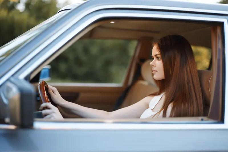 Young woman driving a car with focused expression, natural lighting and green background, showcasing confidence and independence.