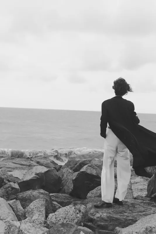 Silhouette of a figure in a long coat with cape, standing on a rocky shore and facing the sea, wind sweeping garments, monochrome scene conveying solitude, contemplation and coastal drama