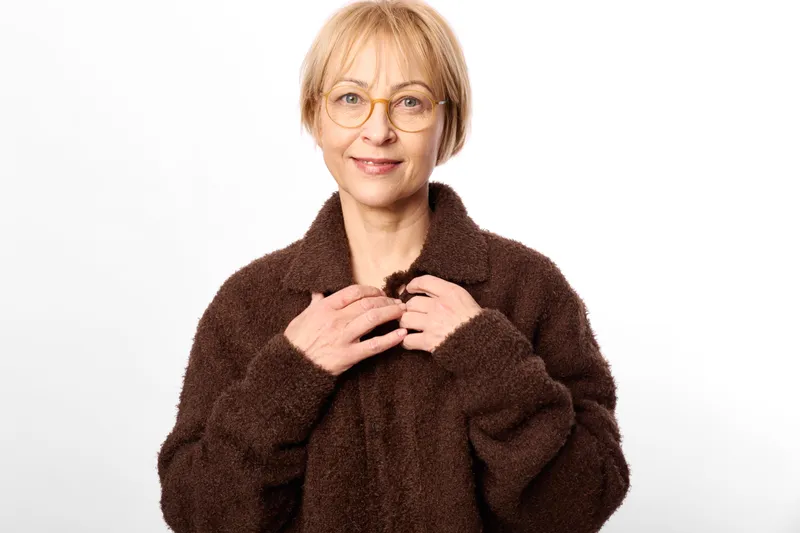 Woman elderly glasses fur coat portrait studio white background, gentle smile, hands on chest, wearing warm brown coat, calm indoor studio lighting, copy space for text only