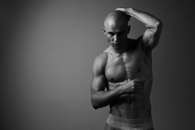 Strong man posing in black and white, showcasing a muscular physique and confident expression. The dramatic lighting highlights his defined features.