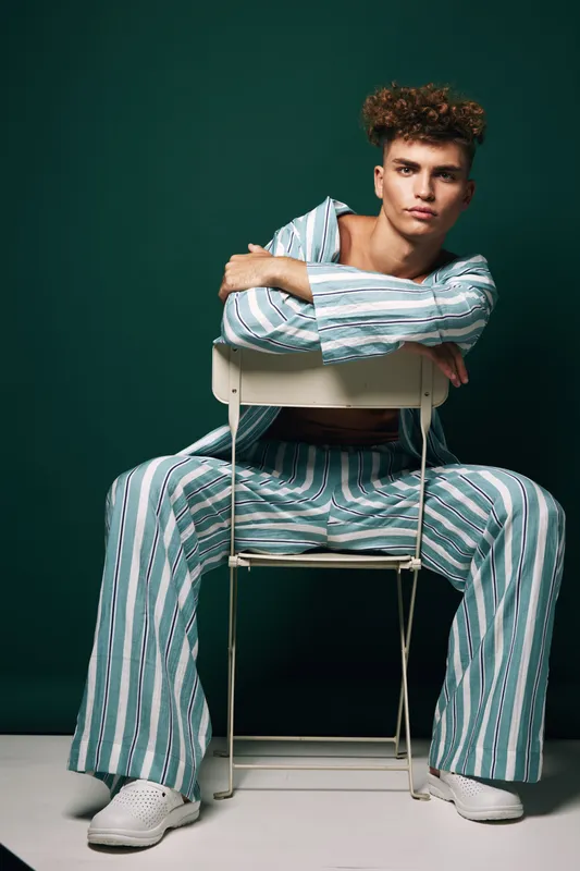 Young man with curly hair wearing striped pajamas sitting on chair backwards against dark green background. Casual fashion, relaxed pose and confident expression in studio setting.