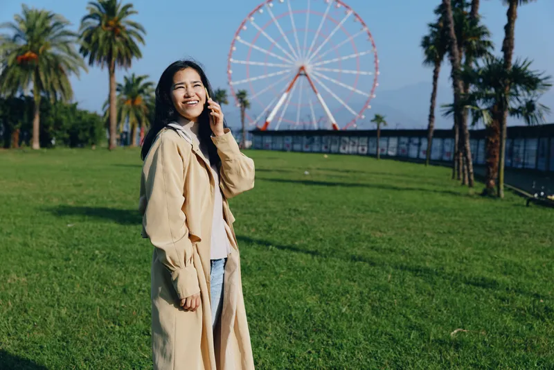 Smiling woman in a long coat stands in a sunny park with a ferris wheel in the background, outdoor leisure scene, casual fashion and joyful atmosphere
