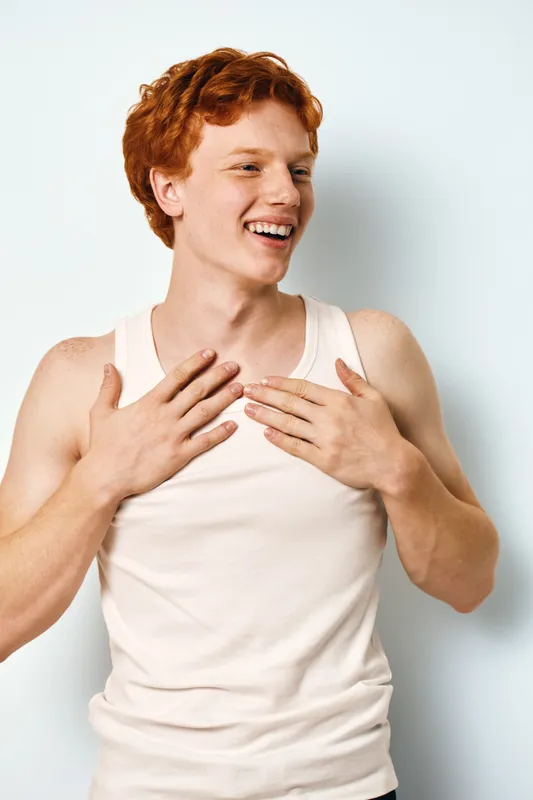 Young man with red hair and freckles wearing white tank top smiling happily with hands on chest against plain white background in casual style portrait.
