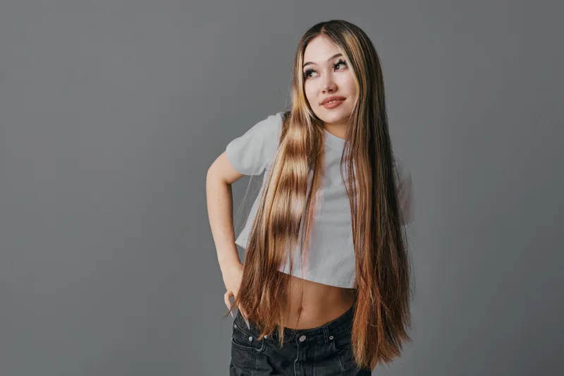 Young woman with long straight hair wearing a white crop top and black jeans posing against gray background looking sideways with hand on hip.