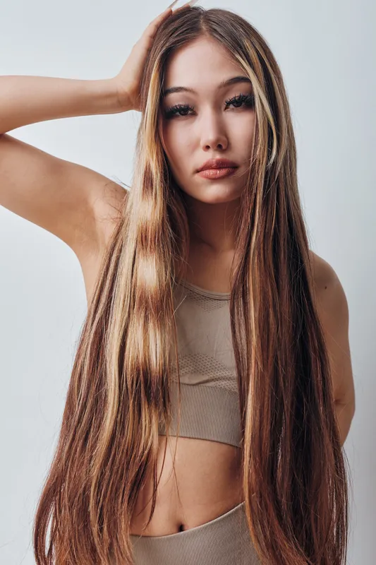 Young woman with long straight hair wearing beige crop top posing in studio. Beauty portrait with natural makeup, confident expression and soft lighting on white background.