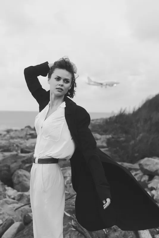 Dynamic coastal fashion shot featuring a wind swept woman wearing a white dress and long coat against rocky shores with distant aircraft