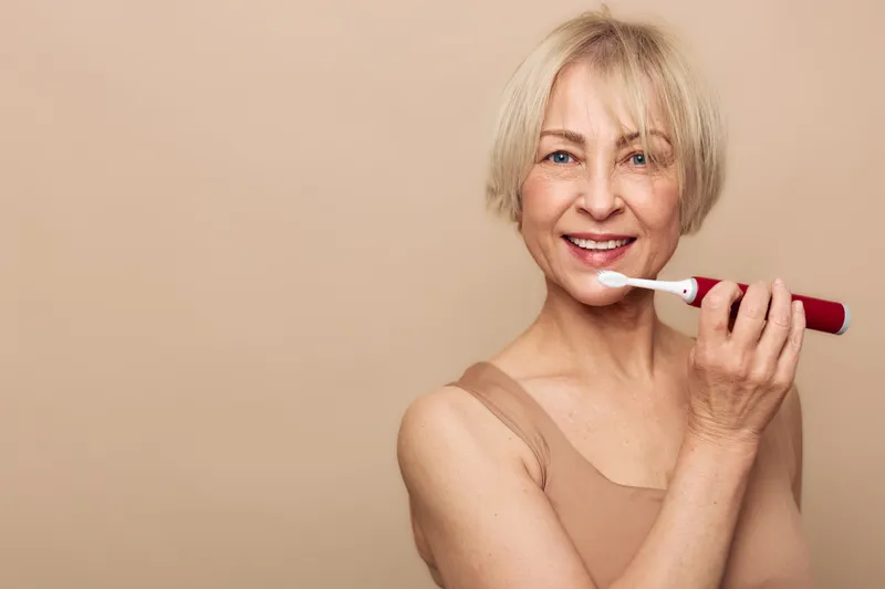 Smiling mature woman brushing teeth with electric toothbrush in beige tank top on beige background. Concept of dental care, oral hygiene, healthy smile, and daily routine for seniors.