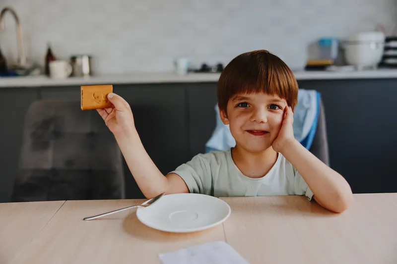 Happy boy holding a cookie, smiling brightly, enjoying a snack at home, showcasing a joyful and playful atmosphere with a cozy kitchen background.