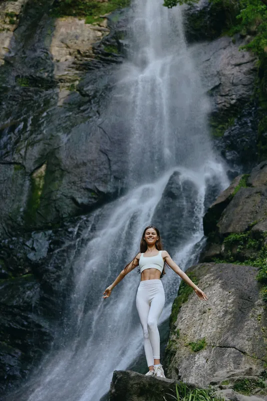 young woman in white activewear stands joyfully in front of a large waterfall surrounded by green mossy rocks and natural scenery during daytime
