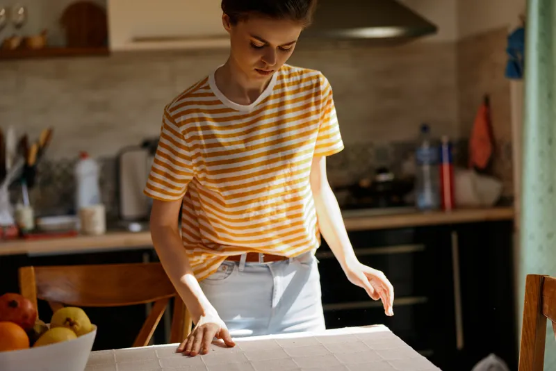 Casual scene of a person in a striped shirt standing at a kitchen island, preparing a task with focus and calm. Natural daylight, soft shadows, home life, and everyday routine.