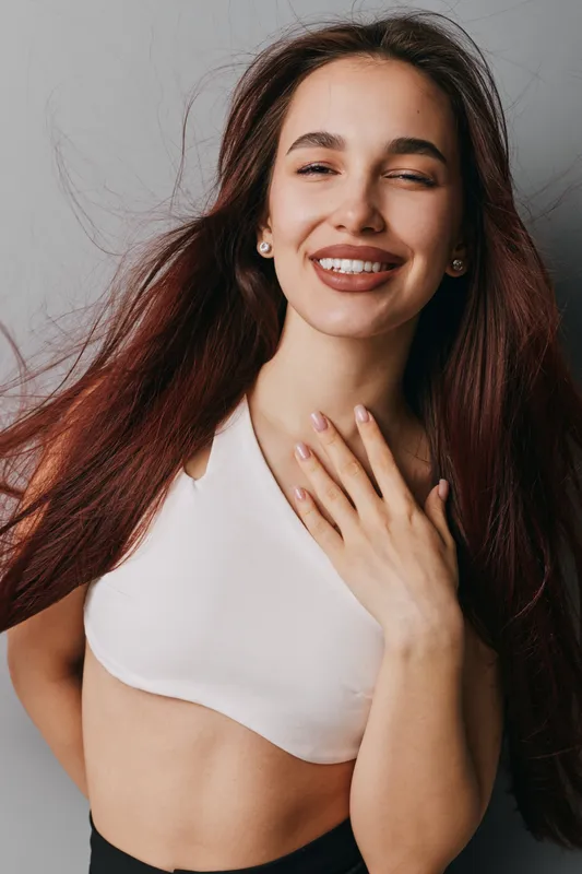 Young woman with long hair smiling with eyes closed and hand on chest wearing white top against neutral background for beauty and happiness concept.