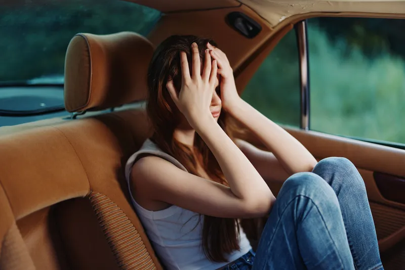 A young woman with long hair, sitting in a car, holding her head, expressing sadness and frustration, with a blurred green background in soft focus.