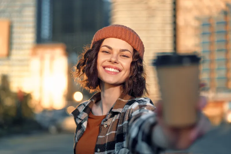 Woman smile coffee city beanie urban portrait outdoors capturing authenticity and candid lifestyle in golden hour glow, mindful living and emotional storytelling with joyful urban warmth.
