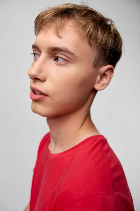 Profile of a young man in a red shirt, calm expression under studio lighting, clean neutral background, fashion study with focused facial features and subtle shadows