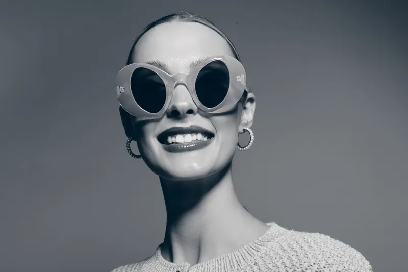 Smiling young white woman with pale skin wearing large round sunglasses and hoop earrings. She wears a knit sweater and poses against a neutral background in studio light.