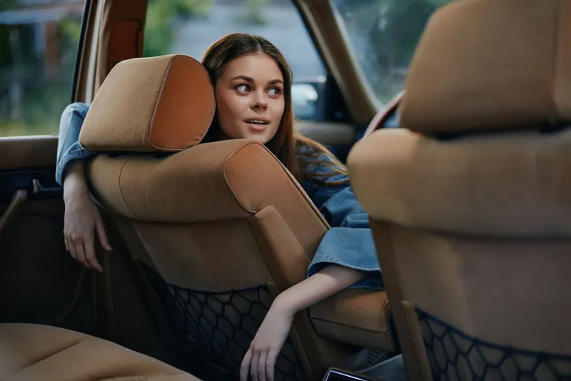 Young female model relaxing in a car's backseat, wearing casual denim clothing, showing a joyful expression against a natural background.