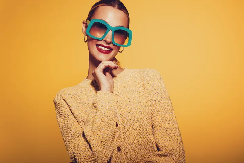 Smiling woman in oversized turquoise sunglasses and beige sweater poses against yellow background. Fashion portrait with bright colors and confident expression. Concept of style and summer mood.