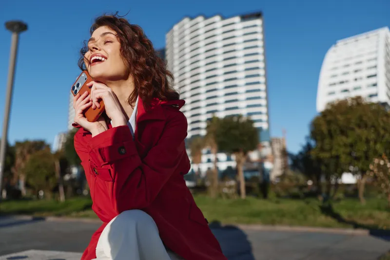 A young woman with curly hair in a red coat smiles while talking on her phone outdoors in a sunny park with modern buildings in the background
