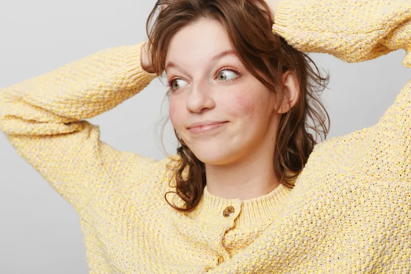 Young woman in yellow knitted sweater looks sideways with playful smile, hands behind head. Casual style, natural light, indoor portrait, relaxed mood, positive expression, cozy clothing.