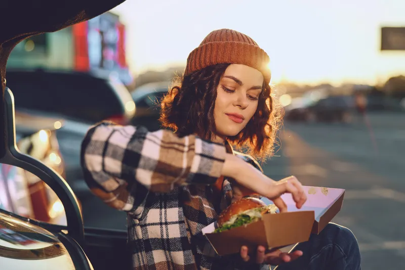 Woman sits at car trunk enjoying a burger from takeout box outdoors at sunset, candid lifestyle and authenticity captured with golden hour glow, mindful living and emotional storytelling.