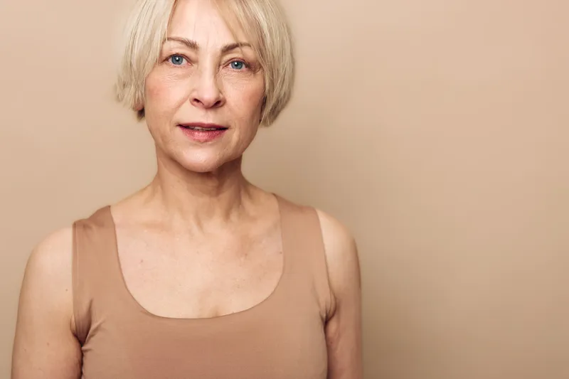 Portrait of a mature woman with short blonde hair wearing a beige tank top. She looks confidently at the camera against a neutral background. Concept of aging and natural beauty.