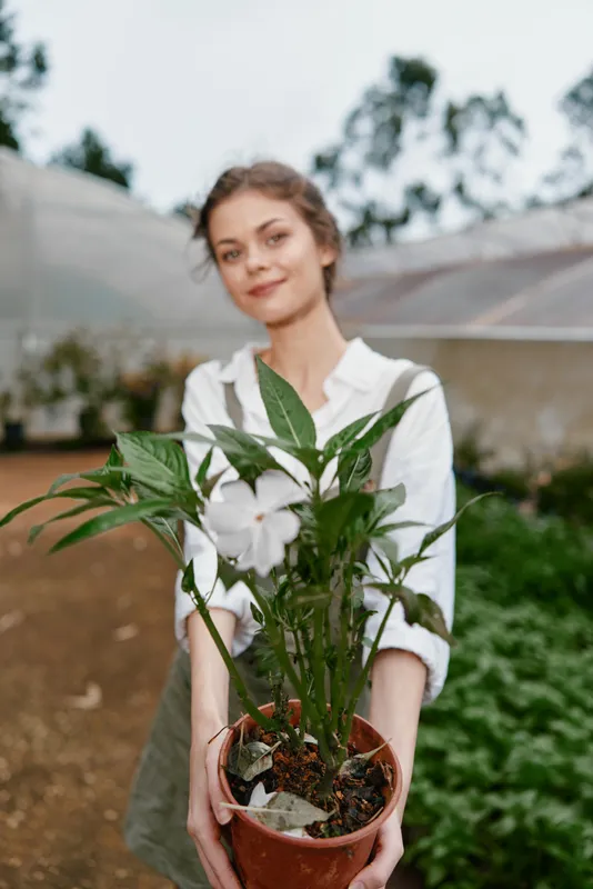 Young woman smiling while holding a potted plant in a greenhouse, wearing a casual white shirt, showcasing the theme of gardening and nature