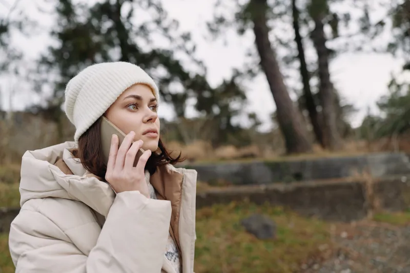 Woman, outdoor, phone, winter, coat, beanie, cold, landscape with trees as she talks on a mobile in a calm chilly park setting during overcast weather
