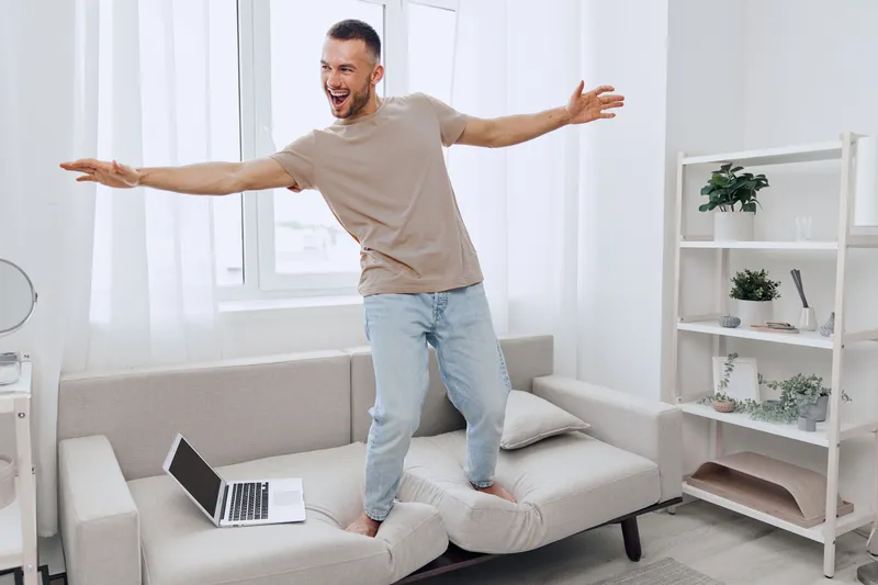 A joyful man enjoying a digital detox while balancing on a couch, symbolizing the blend of work-life and the quest for authentic living, modern lifestyle.