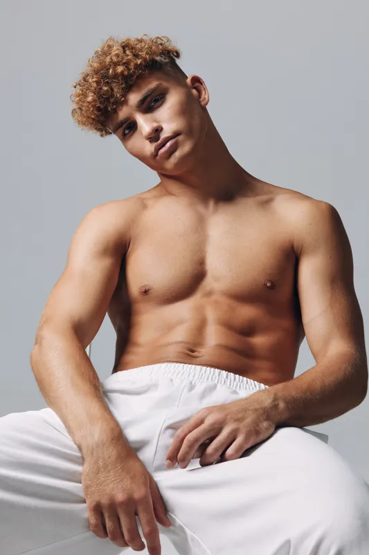 Young man with curly hair and muscular body posing shirtless in white pants against plain background for fitness and fashion concept.