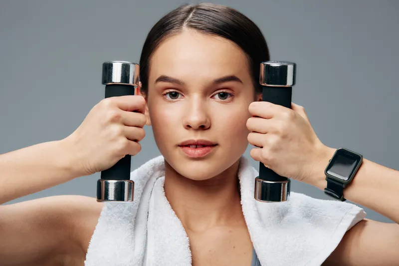 Portrait of a young woman in fitness attire holding two chrome dumbbells near her face, white towel draped on shoulders, smartwatch on wrist, neutral gray studio background.