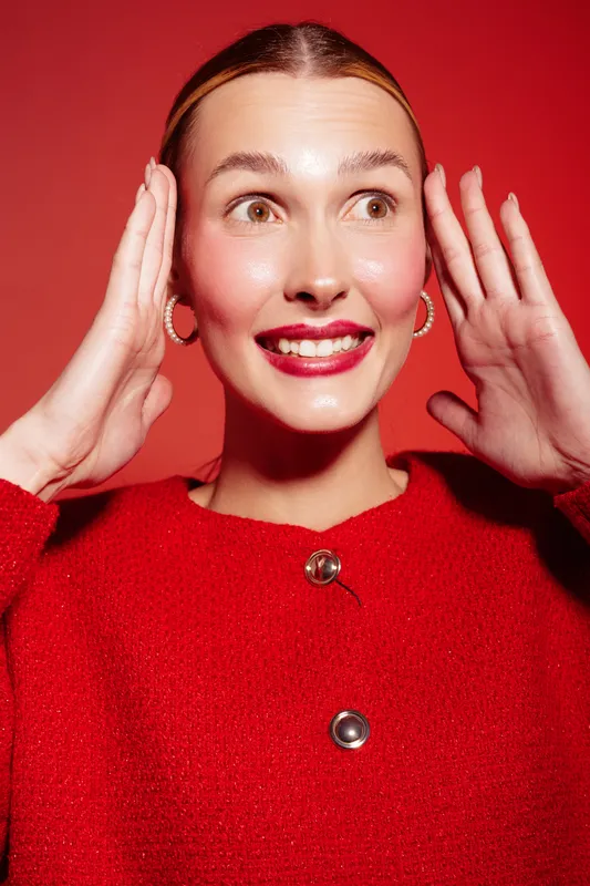 Smiling woman in red sweater with hoop earrings holds hands near face on red background. Bright studio lighting highlights joyful expression and festive mood. Concept of happiness and fashion.