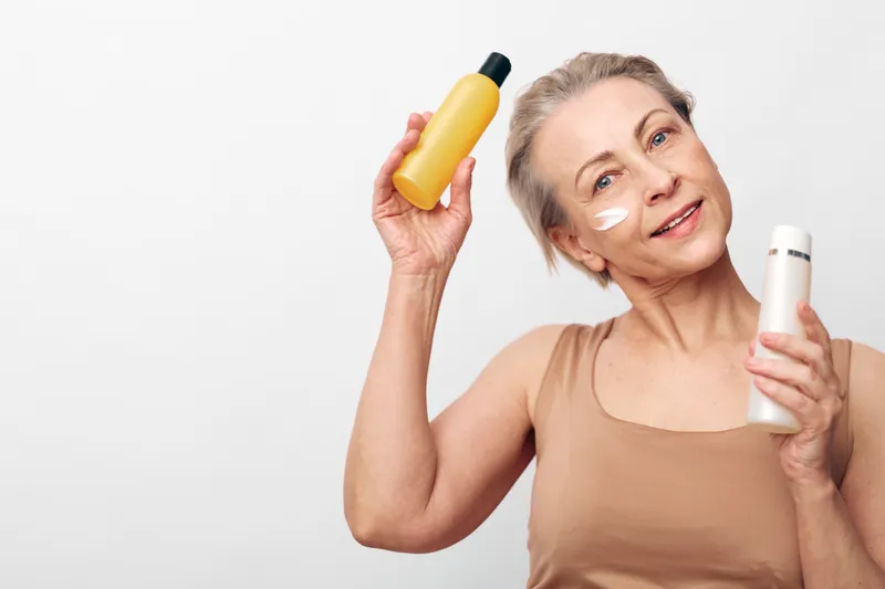 Smiling senior woman with short hair applies face cream holding two skincare bottles in hands. White background, natural beauty concept for aging care and wellness routine.