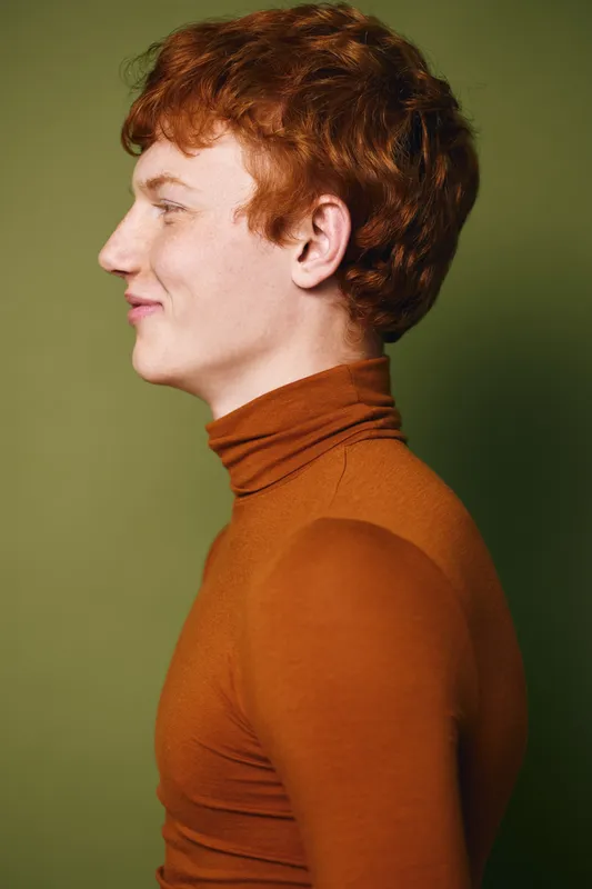 Portrait of smiling young man with red hair wearing orange turtleneck sweater against green background in studio. Profile view of happy person with fair skin and curly hair.