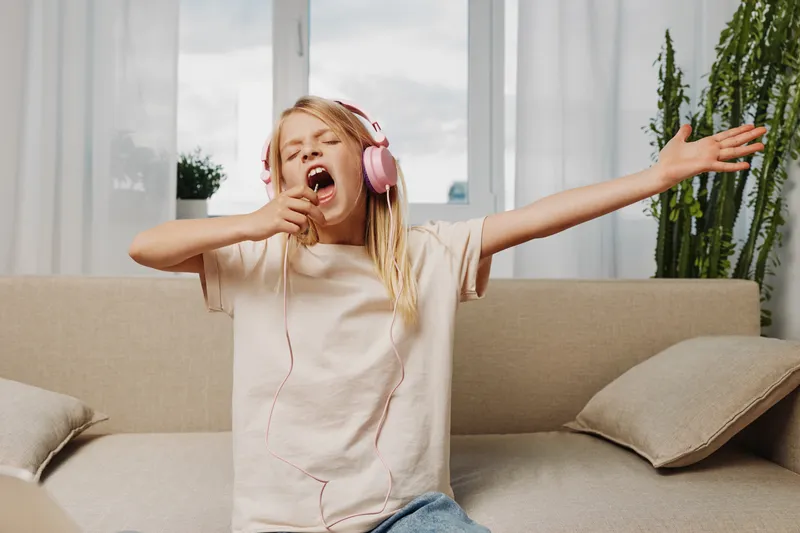 Happy girl with long hair wearing pink headphones sings passionately with eyes closed and hand outstretched sitting on beige sofa in bright living room. Concept of joy, music, and freedom.
