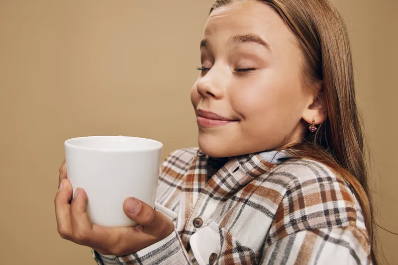 Girl with closed eyes holding white mug enjoying aroma of hot drink in cozy plaid shirt. Relaxation and comfort with warm beverage on beige background.