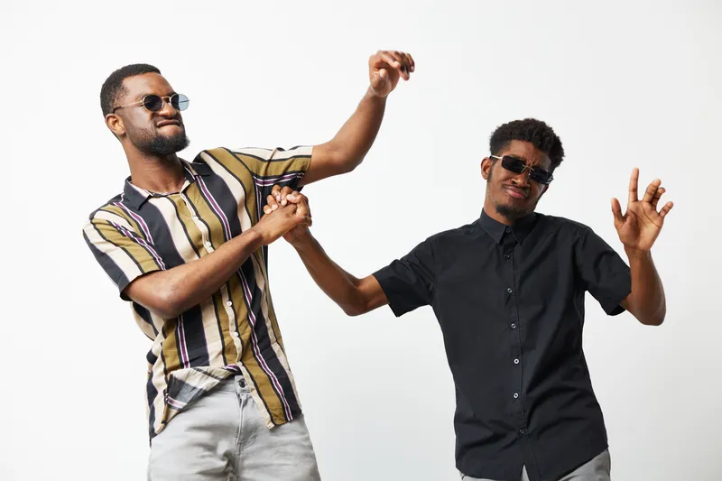 Two joyful African American men dancing together, wearing stylish sunglasses and casual shirts, showcasing friendship and fun in a bright atmosphere.