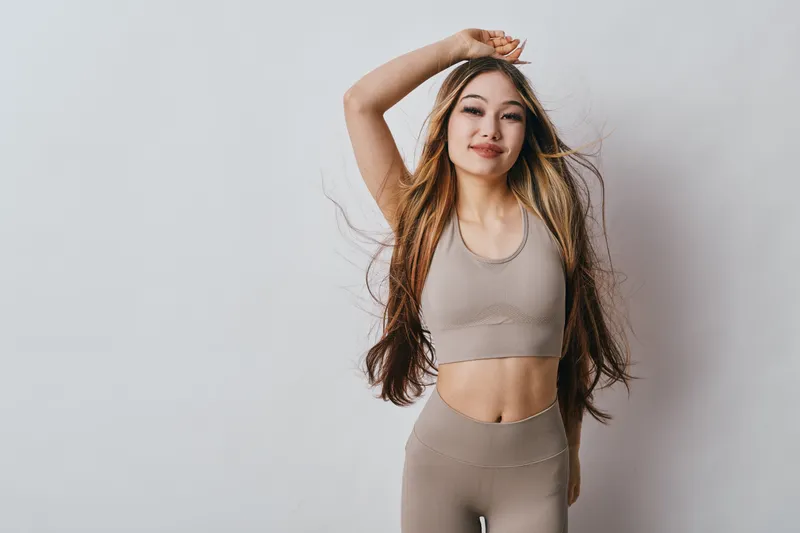 Young woman in beige sportswear posing with hand on head and hair flowing, confident and relaxed against white background for fitness, fashion and lifestyle concept.