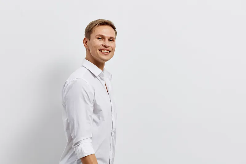 Professional portrait of a cheerful young man with blond hair and blue eyes, dressed in a white shirt, smiling confidently against a plain white background, studio lighting, casual style, happy expres
