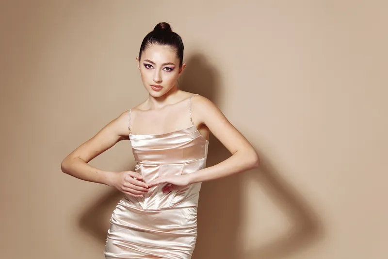 Young woman in a glossy champagne satin dress poses against a beige background in fashion style. Concept combining beauty, elegance, and modern fashion creativity.