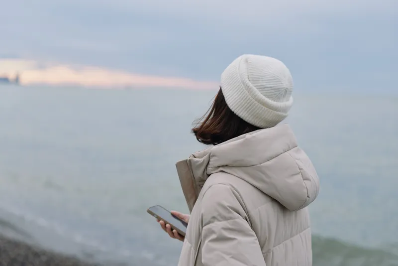 Person in a warm jacket and beanie gazes at a phone along the coast, sea and horizon framed by a tranquil waterfront at dusk, conveying calm contemplation and solitude by the seaside