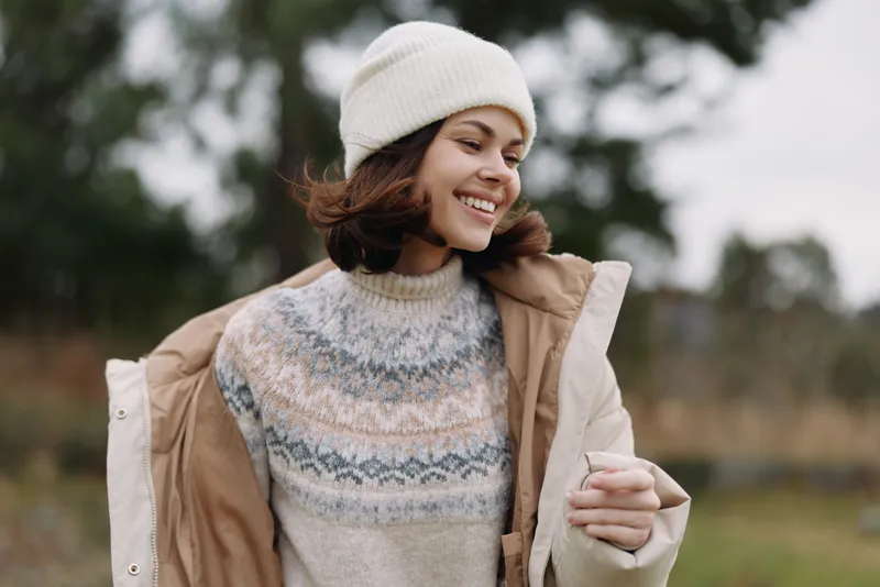 Woman in an outdoor winter scene wearing a knit sweater, warm coat and beanie, smiling as the breeze lifts hair, casual fashion amid a rustic landscape