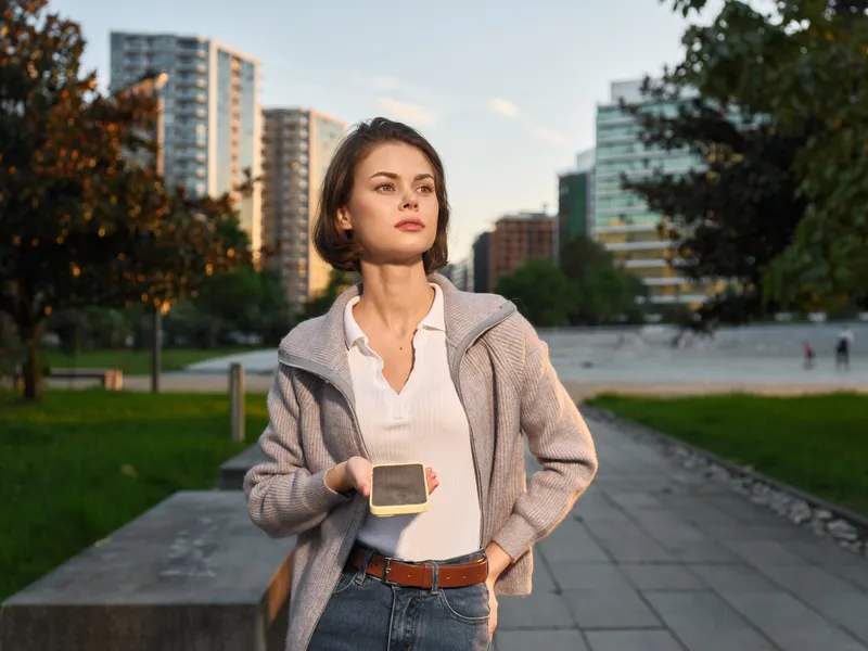 A confident young professional stands outdoors in a modern city park, texting on a smartphone, wearing casual business attire, with a calm, focused expression.