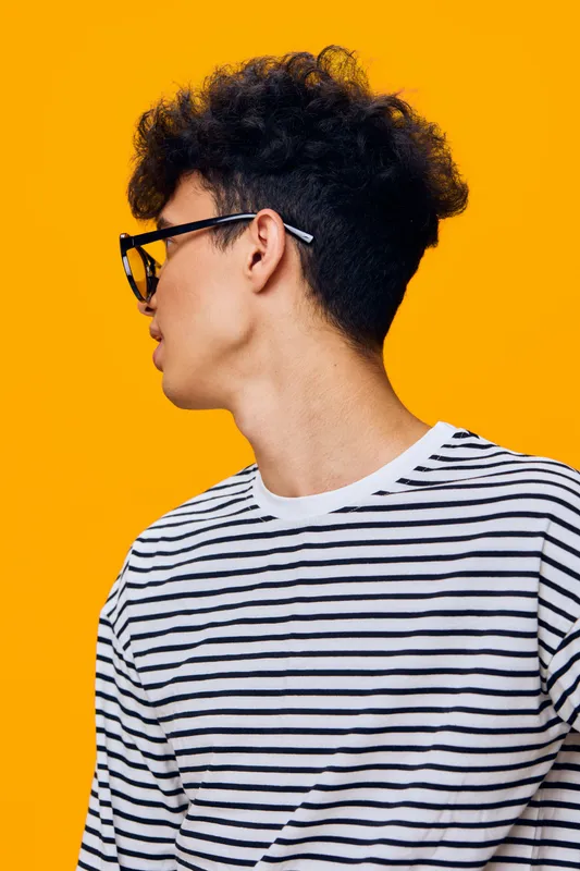 Young man with dark curly hair wearing glasses and a striped shirt looking sideways on bright yellow background, casual style and modern youth fashion.