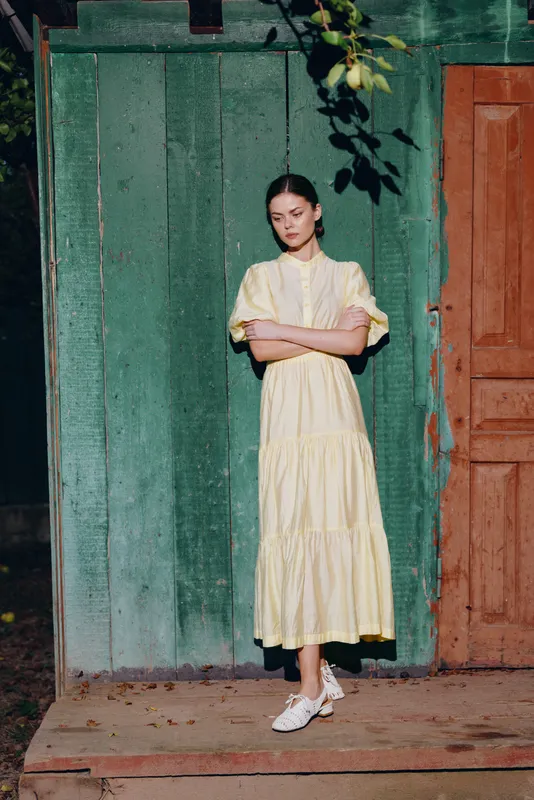 Woman in a pale yellow dress stands in front of a green doorway outdoors, conveying calm elegance and a minimalist fashion portrait in natural light.