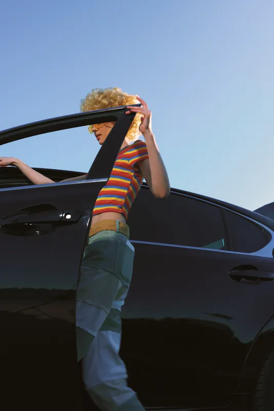 Person stepping from a sleek car under bright blue sky, wearing a colorful striped shirt and jeans, captured in outdoor daylight. Concept of travel, casual fashion, and spontaneity.